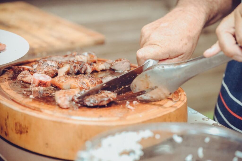 Hand slicing juicy steak on a wooden chopping board with tongs in indoor kitchen.